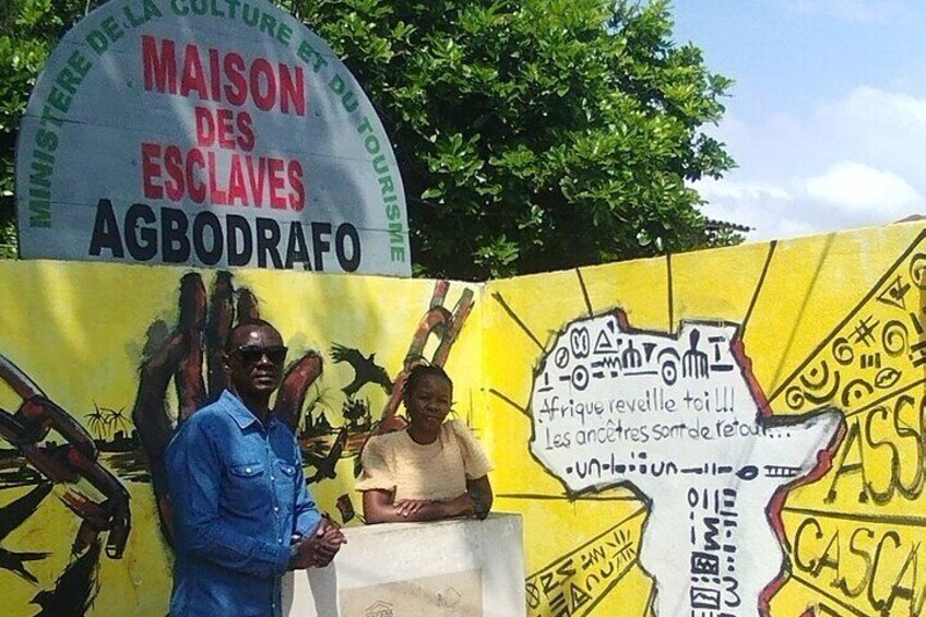Un cartello con la scritta 'MAISON DES ESCLAVES AGBODRAFO' e un murale raffigurante persone e testo. 4
