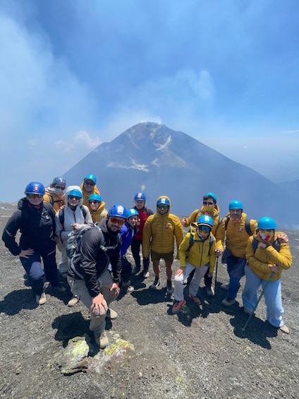 A group of people in outdoor gear posing in front of a mountain with smoke or steam rising from its peak. 3