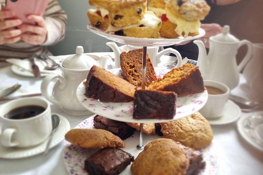 A tiered cake stand with slices of cake and cookies, a teapot, cups, and a saucer on a table. 1