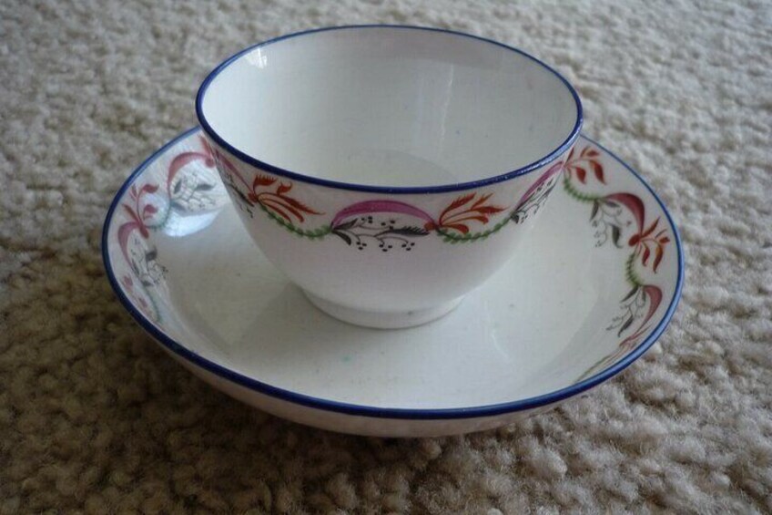 A white teacup with a blue rim and floral pattern, placed on a matching saucer, on a textured carpet. 1