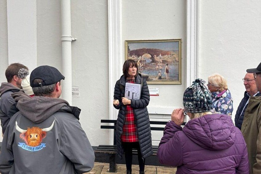 A person presenting to a group outdoors, with a building and a painting in the background. 5