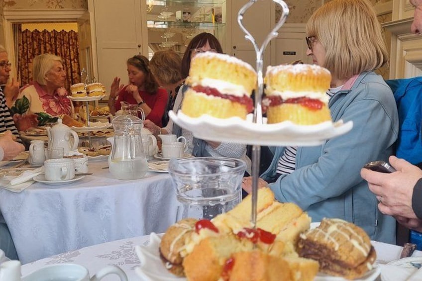 A group of people at a tea party with tea sets, cups, and a variety of pastries on a table. 4