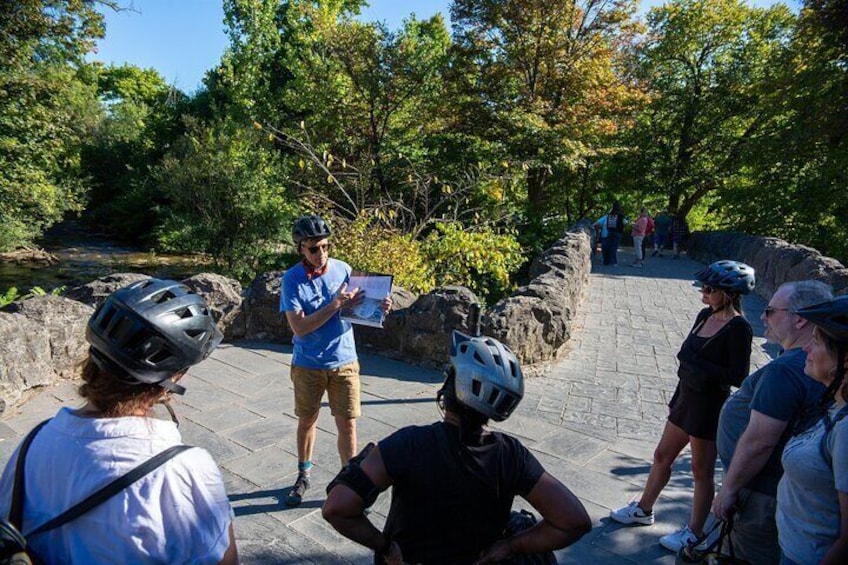A group of people wearing helmets and cycling attire, standing on a paved path in a park with trees and a rocky area in the background. 5