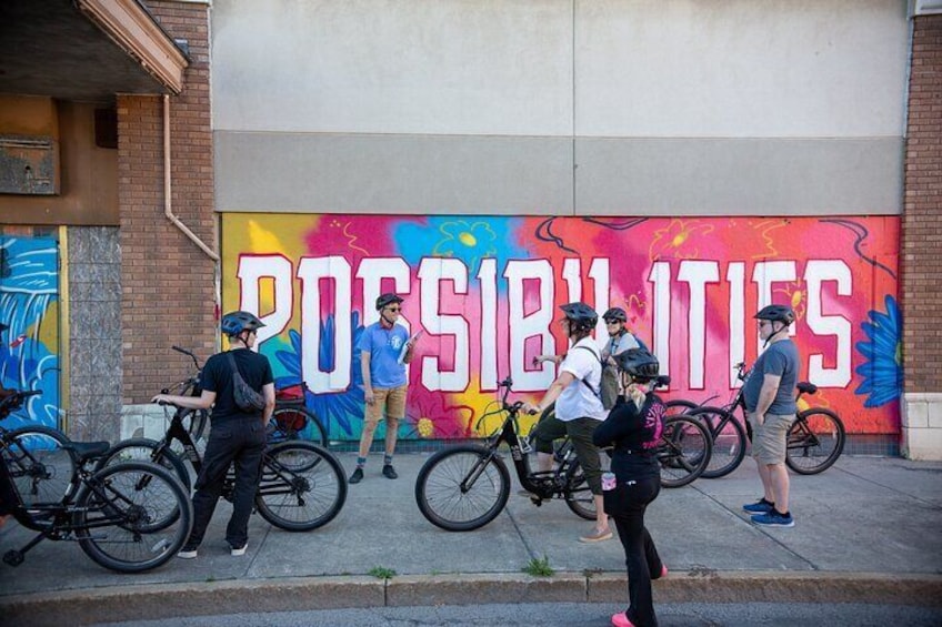 People standing and sitting on a sidewalk in front of a colorful mural with the word "POSSIBILITIES". 3