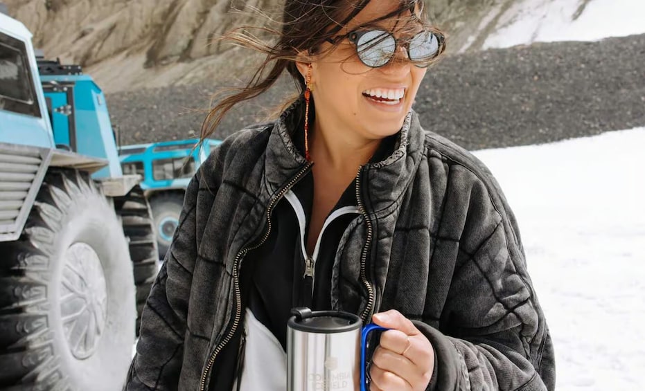 A person holding a stainless steel mug, wearing a dark jacket, with a blue vehicle and rocky terrain in the background. 3