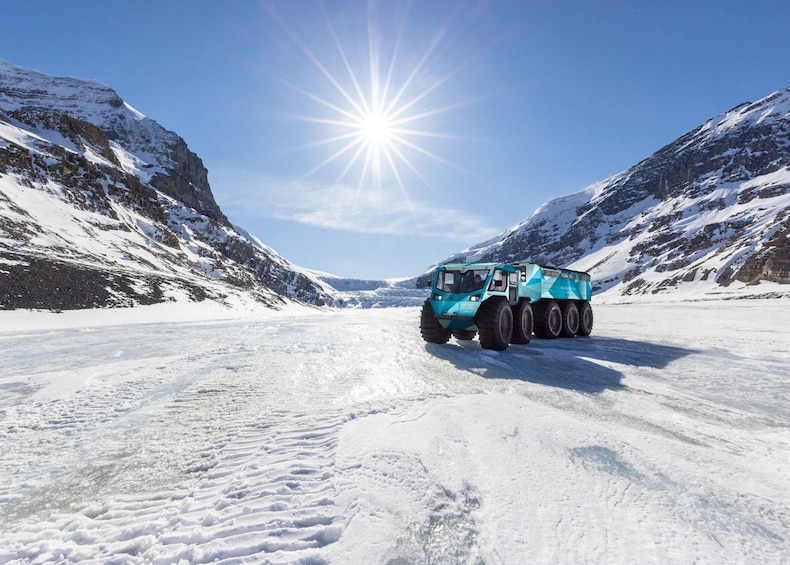 A large blue truck on a snow-covered road with mountains on either side and the sun shining brightly in the sky. 5