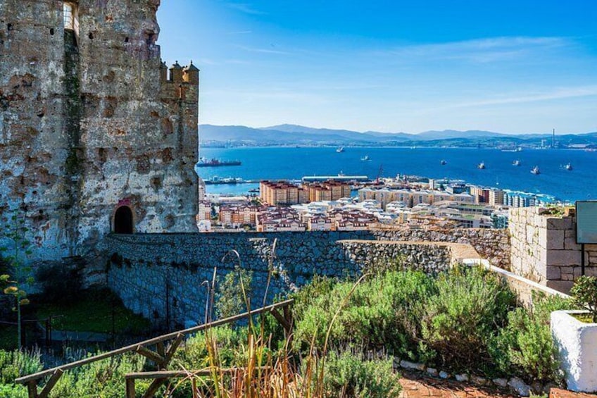 A coastal cityscape with a historic stone tower, a harbor with boats, and a clear blue sky. 5