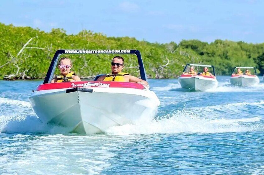 A white boat with a red stripe and the text "Jungle Tour Cancun" on the front. There are two people on the boat, and another boat is visible in the background. 3