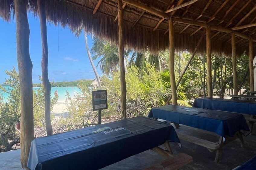 A covered outdoor seating area with tables, a thatched roof, and a view of a beach and palm trees. 2