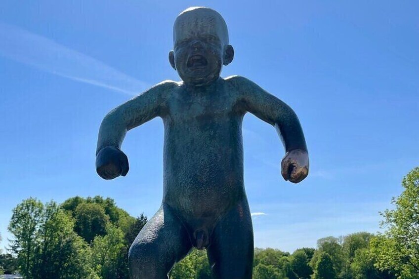 A large bronze statue of a baby with outstretched arms, set against a backdrop of trees and a clear blue sky. 2