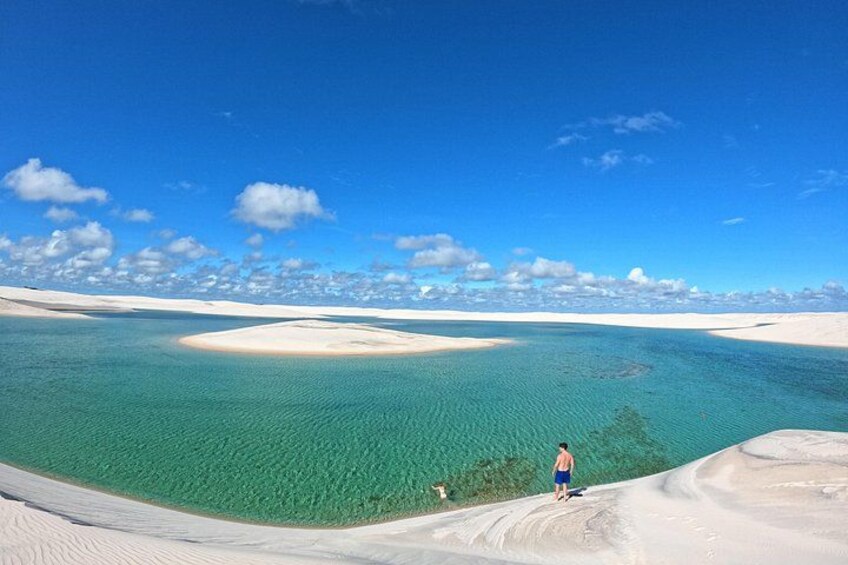 A person standing on a sandy beach with clear turquoise water and white sand dunes in the background. 2