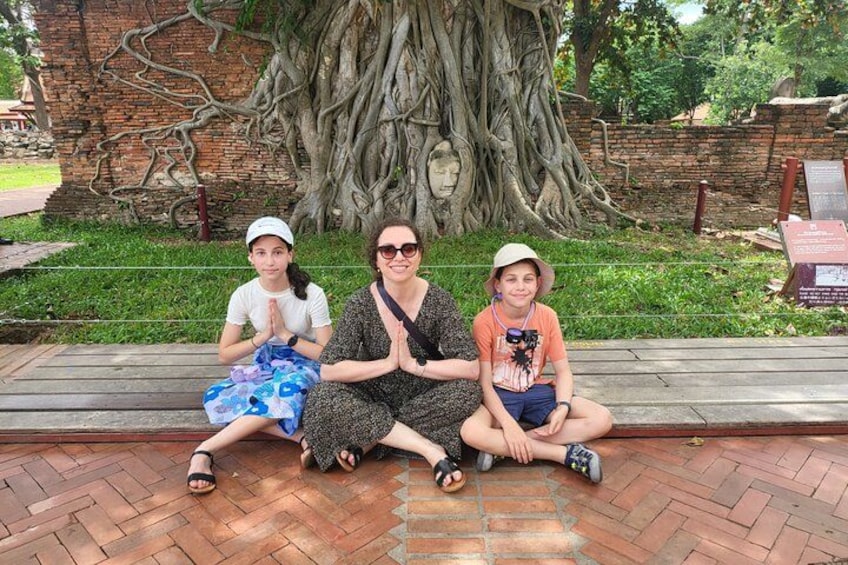 Three people sitting on a wooden bench in a park with a large tree and a brick wall in the background. 4