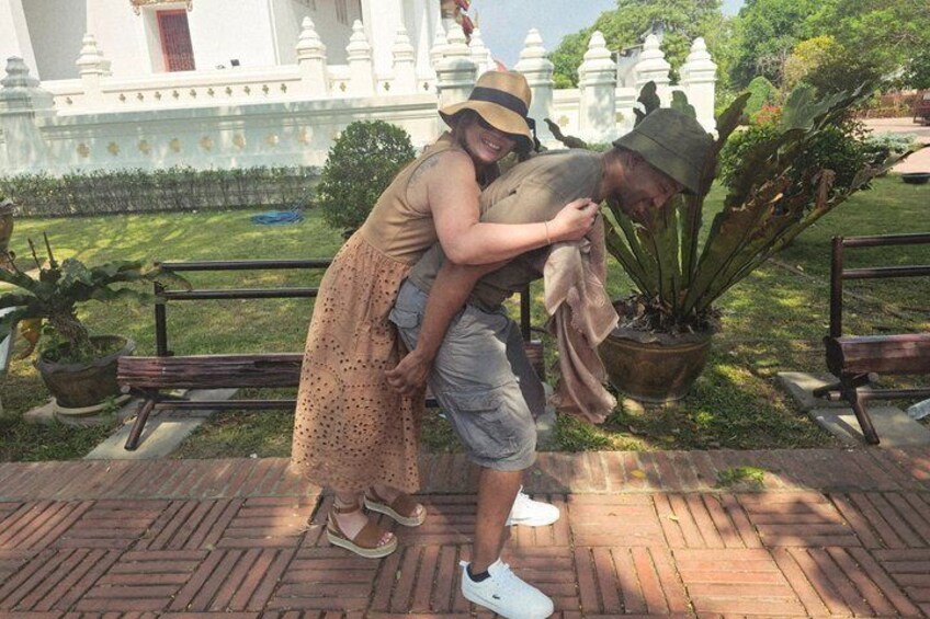 A person in a hat and dress hugging another person in a garden with potted plants and a white building in the background. 4