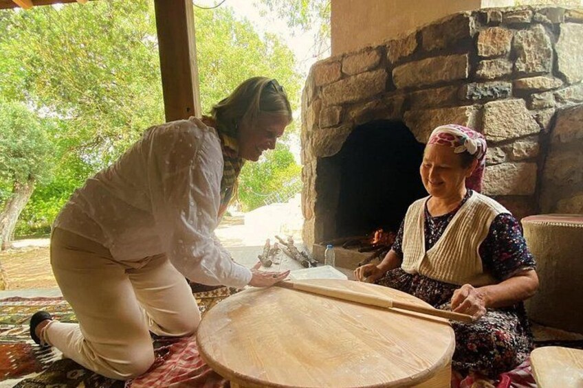 Two people kneeling on a wooden floor, one using a rolling pin on a round wooden surface, and a stone fireplace in the background. 1