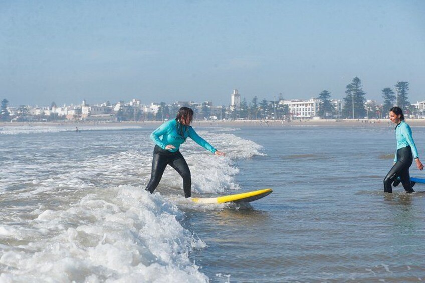 Two people surfing on a beach with waves, a lighthouse, and buildings in the background. 2
