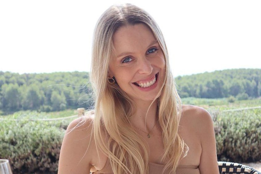 A person with long blonde hair sitting outdoors with a green landscape in the background. 2