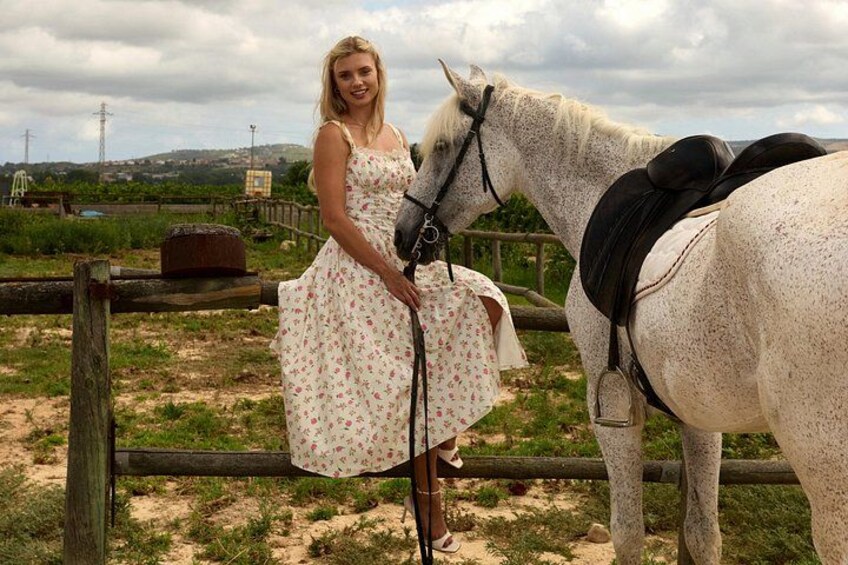 A woman in a floral dress standing next to a white horse with a saddle. 1