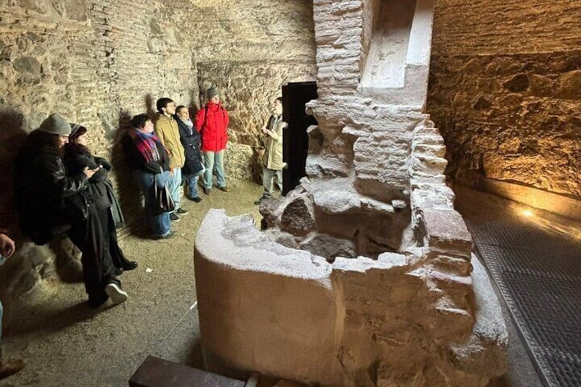 A group of people observing an archaeological excavation site with stone walls and a partially uncovered structure. 1