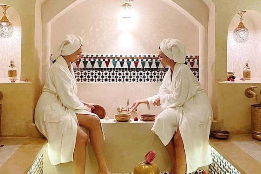 Twee personen in witte gewaden zitten in een traditionele hamam, met een betegelde muur en een klein tafeltje met kommen. 4