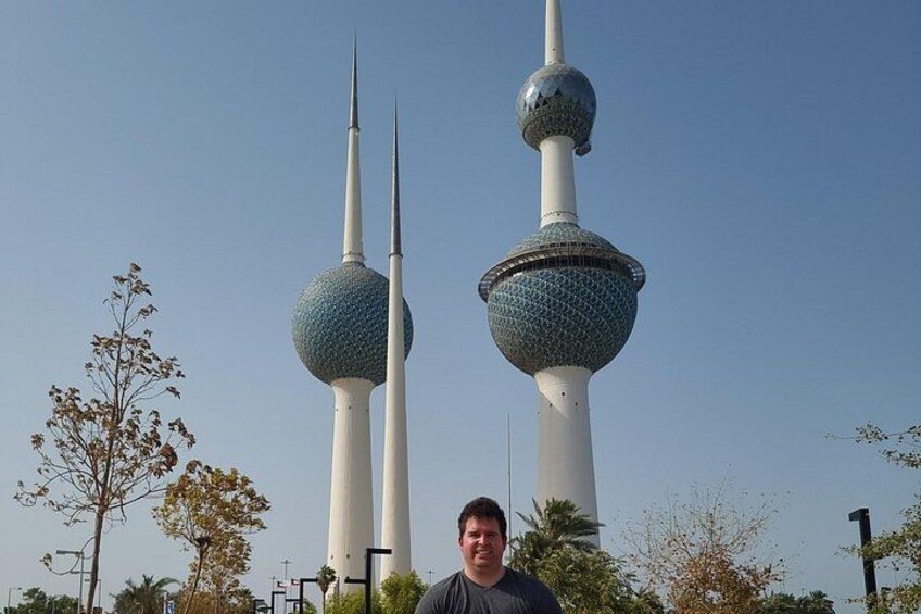 A person standing in front of two tall towers with spherical structures at the top, surrounded by trees and a clear sky. 2