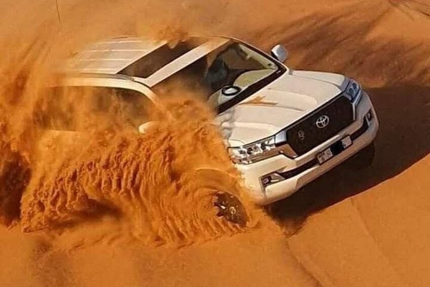 A white vehicle driving through a sandy area with a large amount of sand being displaced. 1