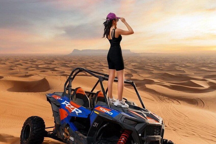 A person standing on a dune buggy in a desert landscape with sand dunes and a distant mountain range. 4