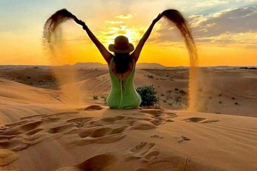 A person in a green dress sitting on sand dunes with arms raised, facing a sunset. 3