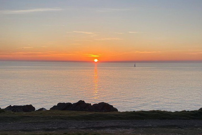 A sunset over the ocean with a sailboat in the distance and rocky outcrops in the foreground. 2