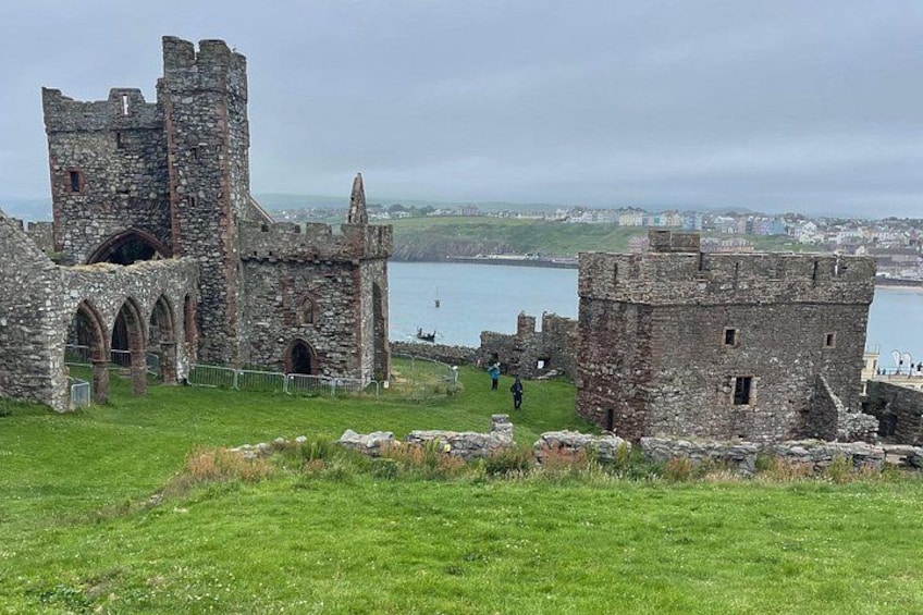 A historic stone castle with towers and arches, a grassy foreground, and a body of water in the background. 3