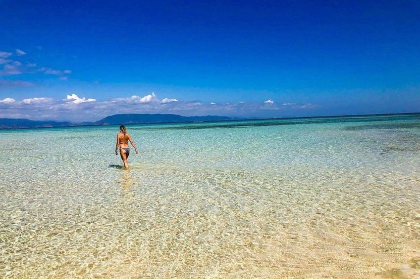 A person walking on a beach with clear water and distant mountains. 4