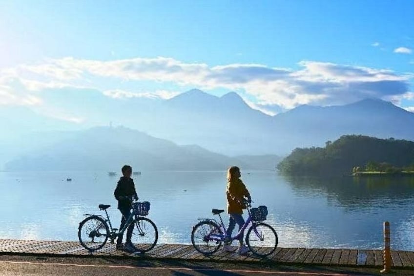 Two people on bicycles by a calm lake with mountains in the background. 1