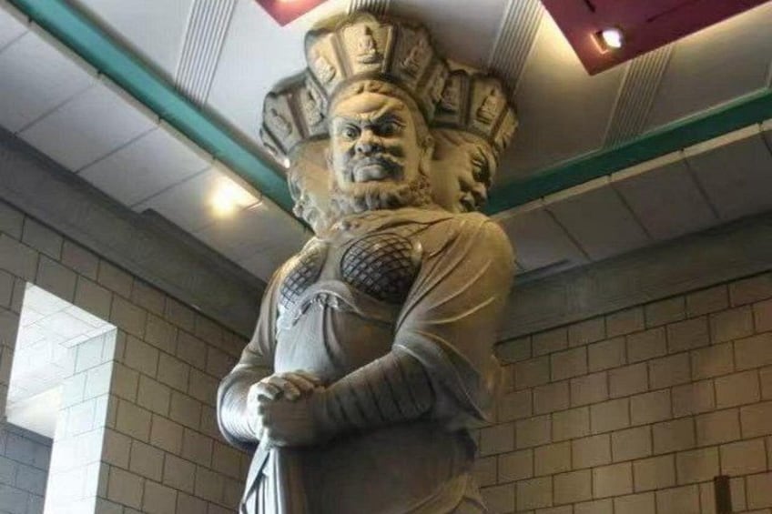 A large statue with a detailed headdress and armor, standing in an indoor setting with a tiled wall and a ceiling with recessed lighting. 2