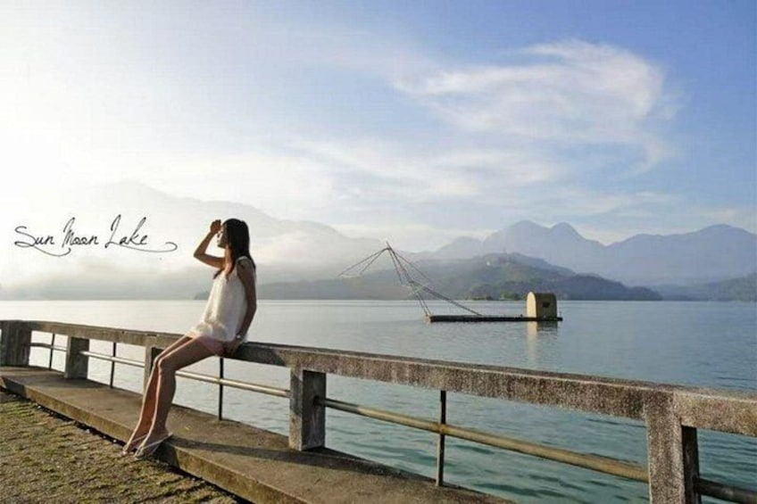 A person sitting on a concrete ledge by a lake with mountains in the background and a bridge in the distance. 5