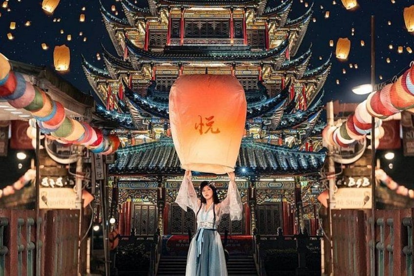 A person holding a lantern with Chinese characters, standing in front of a traditional Chinese building with multiple tiers and ornate decorations. 1