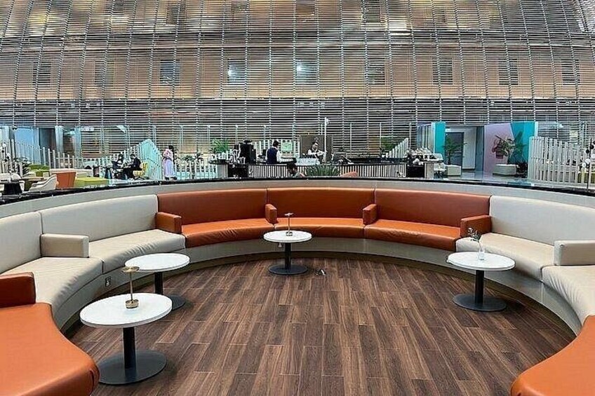 Un ambiente interno moderno con una zona salotto dalle linee curve, tavoli rotondi e un soffitto alto caratterizzato da una struttura a griglia. 1