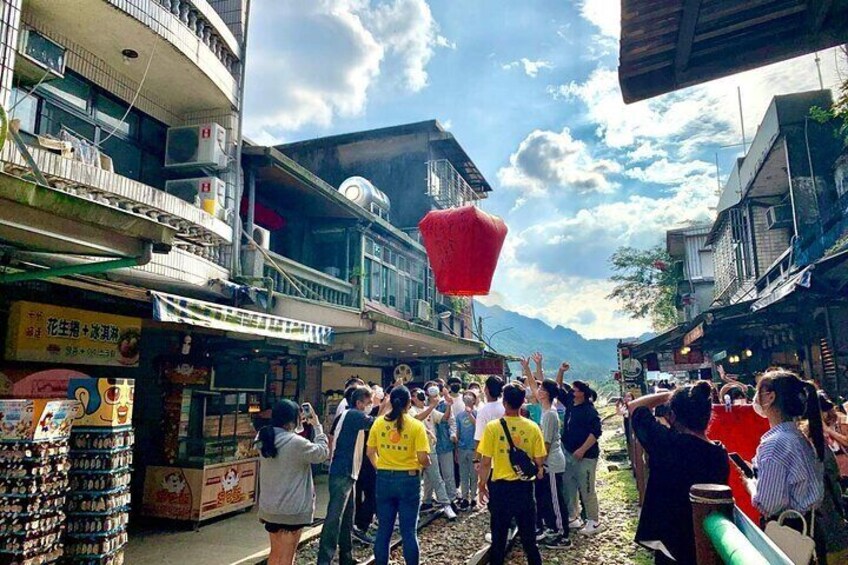 A crowd of people gathered in an outdoor market area with buildings and a mountainous backdrop. 3