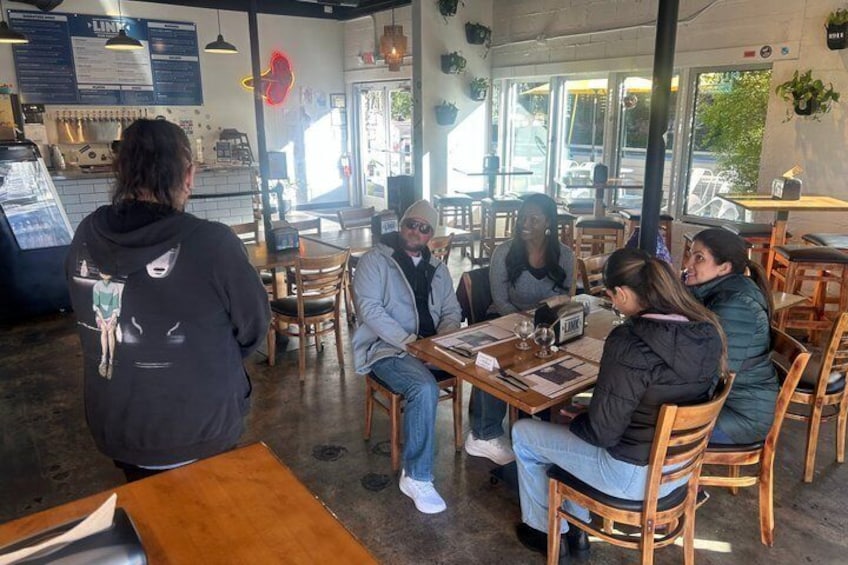 A group of people seated at a table inside a cafe with wooden chairs and tables, a menu board, and a counter with a display. 4