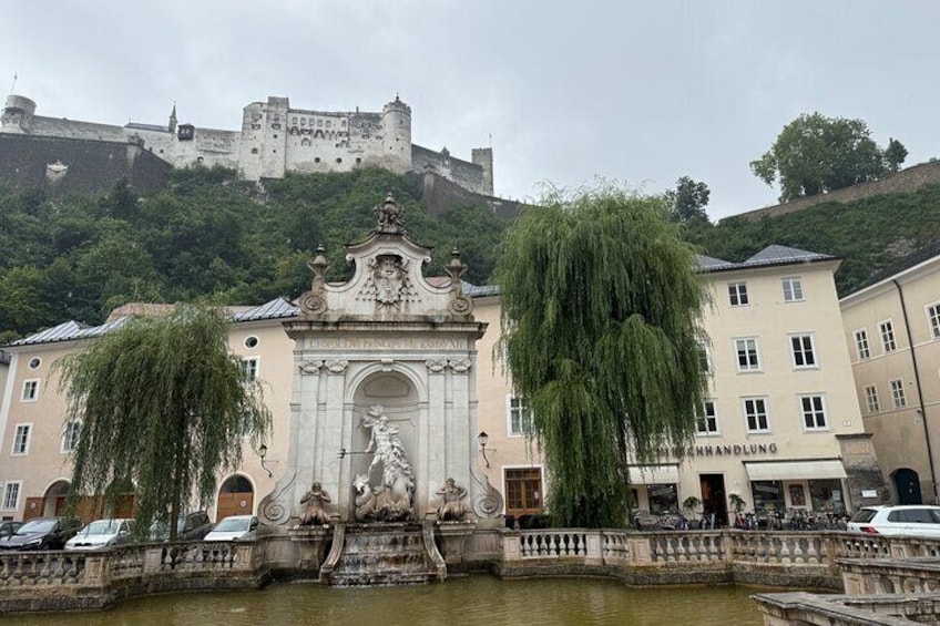 A historic building with a statue in front, a castle in the background, and a river in the foreground. 1