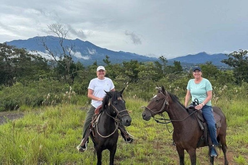 Two people riding horses in a grassy field with mountains in the background. 3