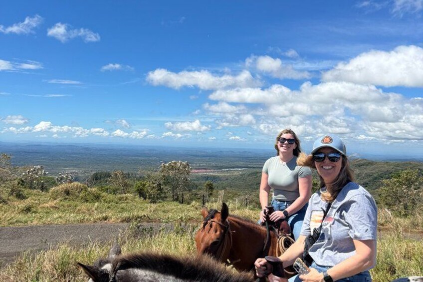 Two people sitting on a grassy hill with a horse, overlooking a valley. 3