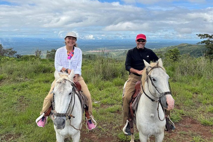 Two people riding horses on a grassy hill with a view of the ocean in the background. 5