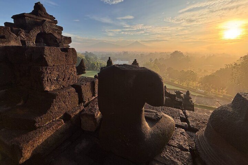 Vestiges d’une ancienne construction en pierre, avec une grande sculpture représentant une tête, le tout se détachant sur le fond d’un coucher de soleil sur un paysage parsemé d’arbres et de collines lointaines. 4