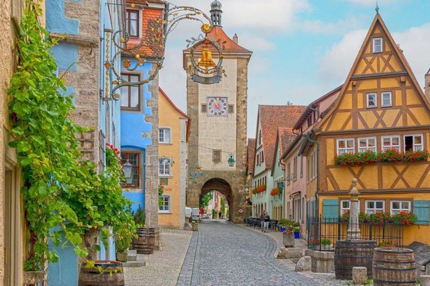 A cobblestone street with colorful half-timbered houses, a clock tower, and a wrought iron gate. 1