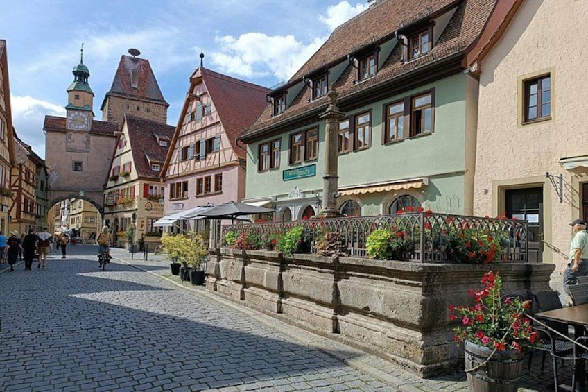 A cobblestone street lined with traditional half-timbered houses, a stone bridge, and a clock tower in the background. 2