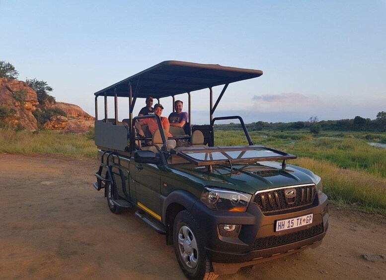 A safari vehicle with a roof, two passengers, and a license plate 'HH 15 TX GP' on a dirt road with grass and rocks in the background. 3
