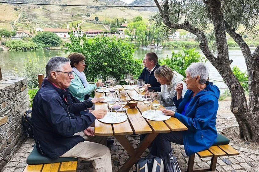 A group of people dining outdoors at a wooden table with a scenic view of a river and hills in the background. 5