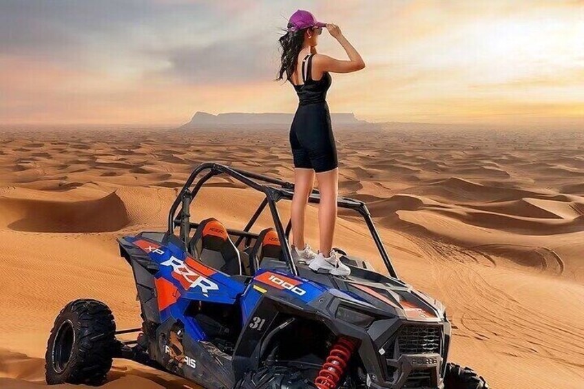 A person in a black outfit standing on a blue and orange off-road vehicle in a desert with sand dunes. 1