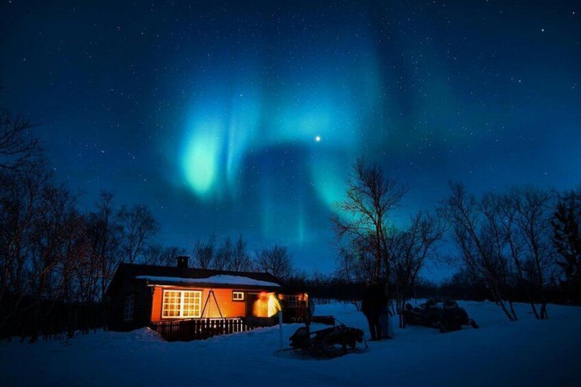 A house with a lit window and a snow-covered landscape under a starry night sky with the aurora borealis. 3