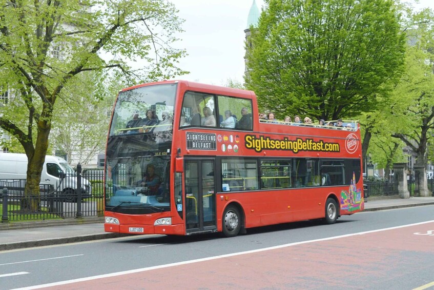A red double-decker sightseeing bus with passengers on the upper deck, driving on a city street lined with trees and a building in the background. 1