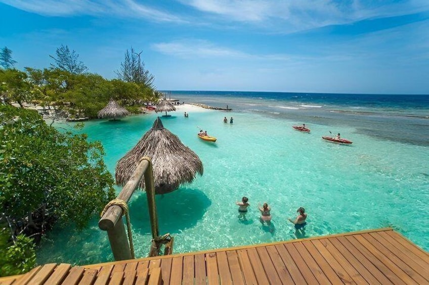 A wooden deck overlooking turquoise waters, a thatched-roof hut, and people swimming and kayaking. 5